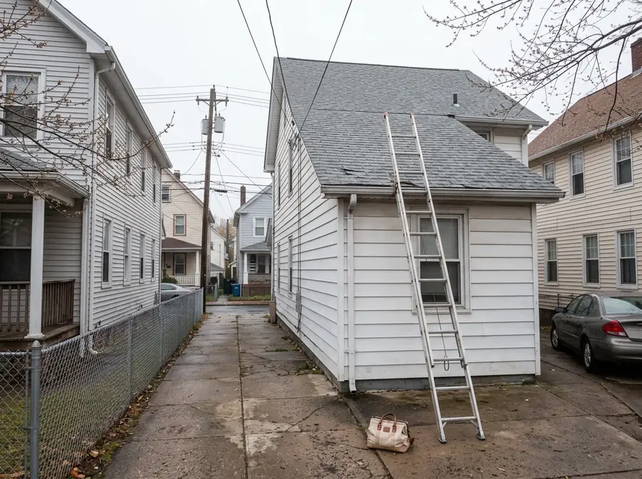 Ladder leaning against the roof edge of a white frame house beside a narrow driveway