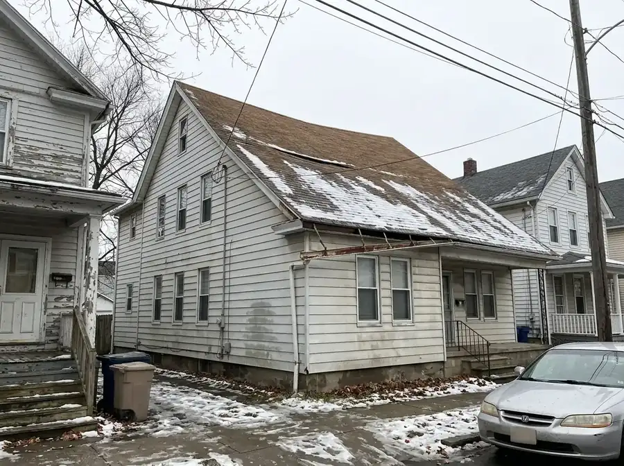 Older two-family house with a dipping brown shingle roof and light snow on a narrow street