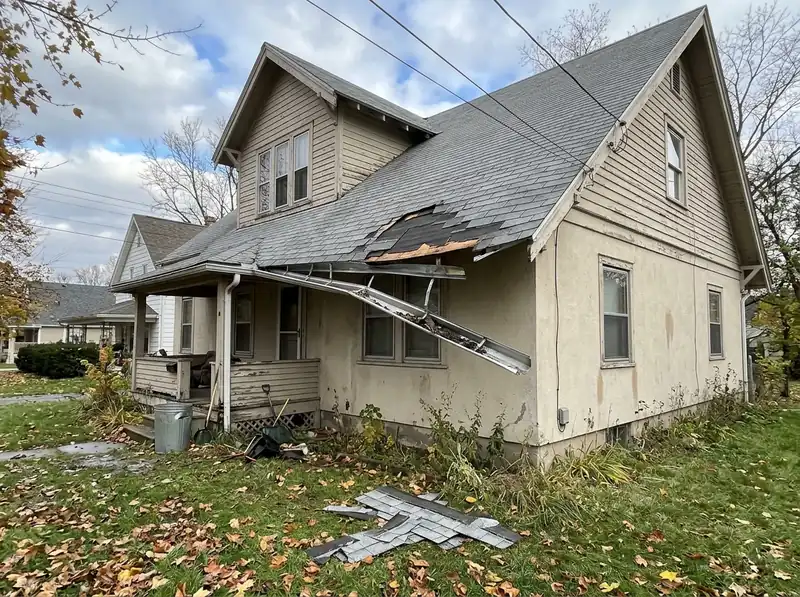 Torn shingles, exposed decking and a hanging gutter on a house, with shingles on the lawn